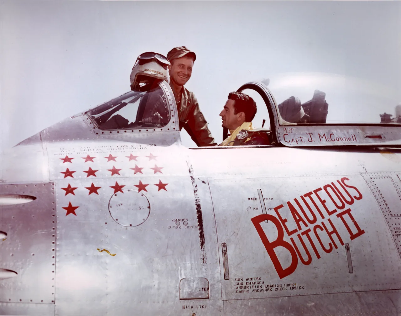 A pilot in flight gear sits in the open cockpit of a silver F-86 Sabre jet while a second man stands beside it. Red stars and the name Beauteous Butch II are painted on the fuselage.