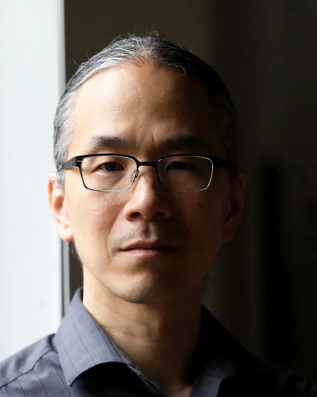 Portrait of Ted Chiang, a man with grey hair tied back and glasses, standing beside a window.