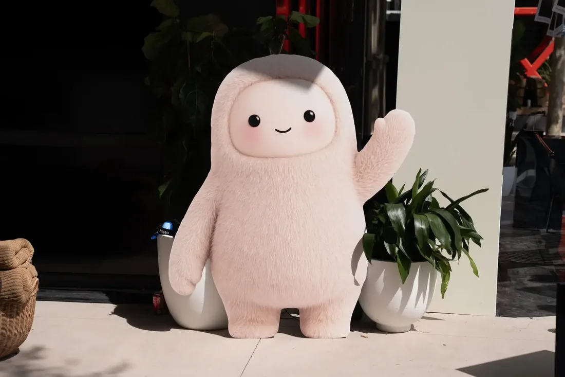 A person-sized mascot covered in pale pink fur, with a round face, two dot eyes and a small smile, waving outside a building.